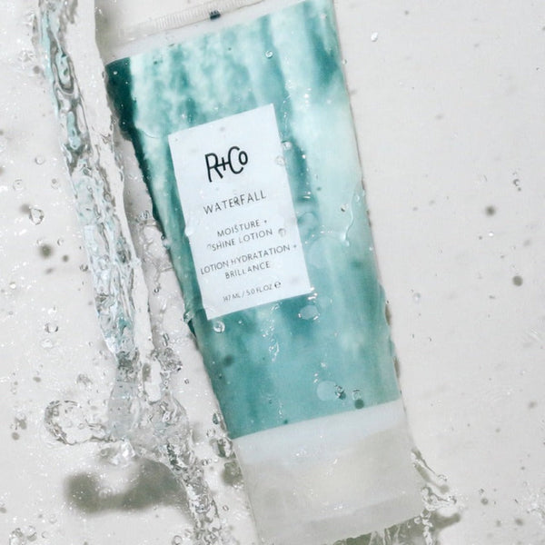 a tube of R+Co Waterfall Moisture + Shine Lotion under a stream of water