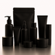 A set of matte black cosmetic and skincare containers, including pump bottles, dropper bottles, tubes, jars, and a stand-up pouch, arranged on a reflective surface against a clean white background.