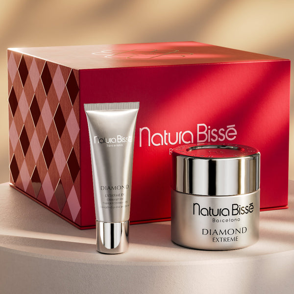 NaturBissé skincare set featuring a red gift box, a silver shampoo tube, and a silver cream jar on a beige pedestal against a warm beige background with soft shadows.