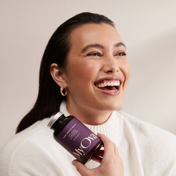 A woman in a cream knit sweater holds a purple bottle of MyOva dietary supplements, smiling and looking off-camera against a warm beige background.