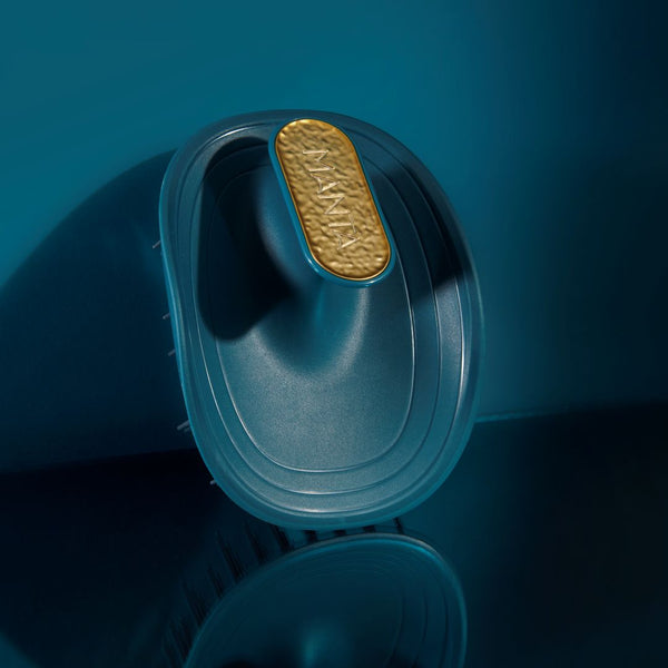 A close-up studio shot of a dark teal hairbrush with a textured gold oval detail on the handle, set against a matching dark teal background.