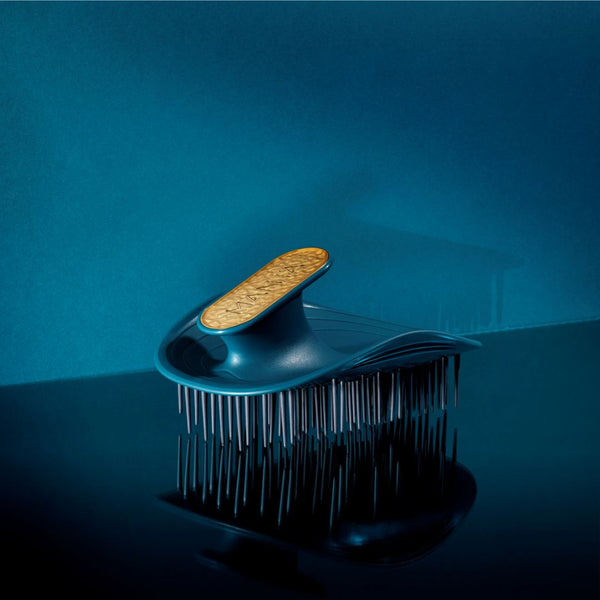 A modern hairbrush with a dark teal-blue body, silver metal bristles, and a cork-textured gold handle, set against a dark teal-blue background.