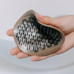A hand holding a metallic bronze hairbrush with black bristles, set against a soft, out-of-focus white background.