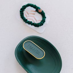 A dark green silicone hair oil drop set by Manta, featuring a small oval bottle with a gold-trimmed cap and a matching dark green silicone hair oil drop, displayed on a light speckled surface.