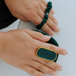 A close-up of hands wearing a dark green velvet ring with a gold border and a small American flag charm, alongside a matching dark green scrunchie on the other hand.