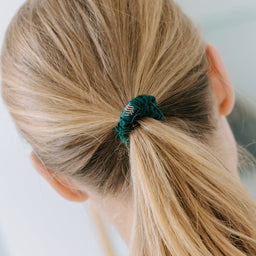 A close-up back view of a person with blonde hair tied back in a low ponytail using a dark green textured scrunchie.