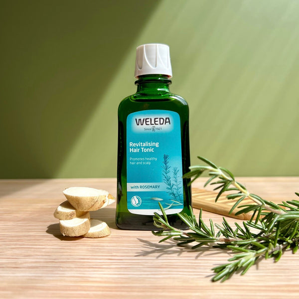 Weleda bottle of revitalizing hair tonic on a wooden surface with rosemary leaves and bulbs.