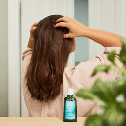 Person with hands in hair next to a bottle of shampoo on a table with plants in the background