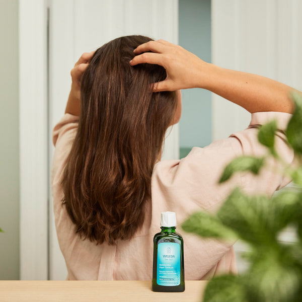 Person with hands in hair next to a bottle of shampoo on a table with plants in the background