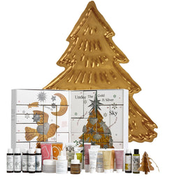 An advent calendar set from Klyve, featuring a large gold Christmas tree-shaped box behind a white advent calendar with gold illustrations, surrounded by various skincare and body care bottles and tubes.