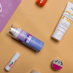 Cosmetic products including a spray bottle, tube, and small containers on a beige surface with a purple leaflet.