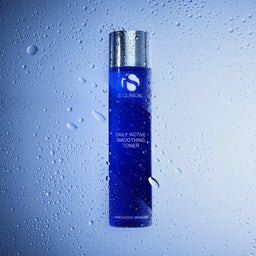 A bottle of e:CLINICAL Daily Active Smoothing Toner with a metallic silver cap and a deep blue body, shown against a cool blue gradient background with water droplets on a glass-like surface.