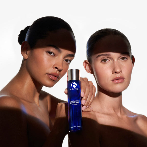 Two models with dark skin and black head wraps pose together while holding a blue bottle of Aesop Daily Active Clay Mask Cleanser against a white background.