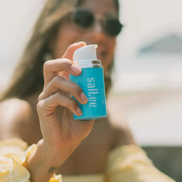 an out of focus woman holding a bottle of Saltee SPF50 Face Daily Protection Formula to the camera