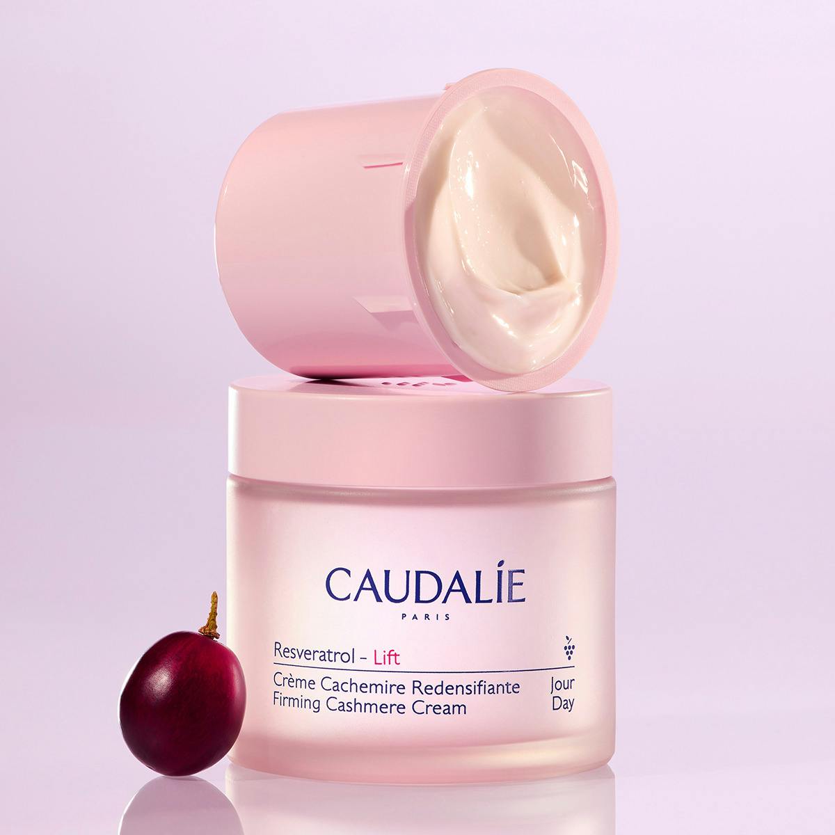 Caudalie Resveratrol - Lift Firming Cashmere Cream jar in pale pink, with the lid off and cream visible, alongside a dark red cranberry on a light lavender background.