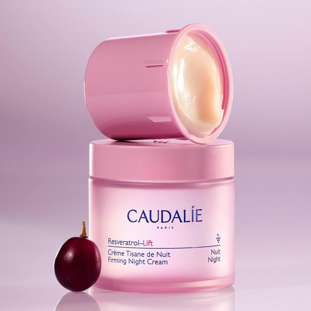 Caudalie Resveratrol-Lift Crème Tisane de Nuit Firming Night Cream in a pink jar, with the lid resting open on top and a dark red resveratrol berry placed beside it on a soft lavender background.
