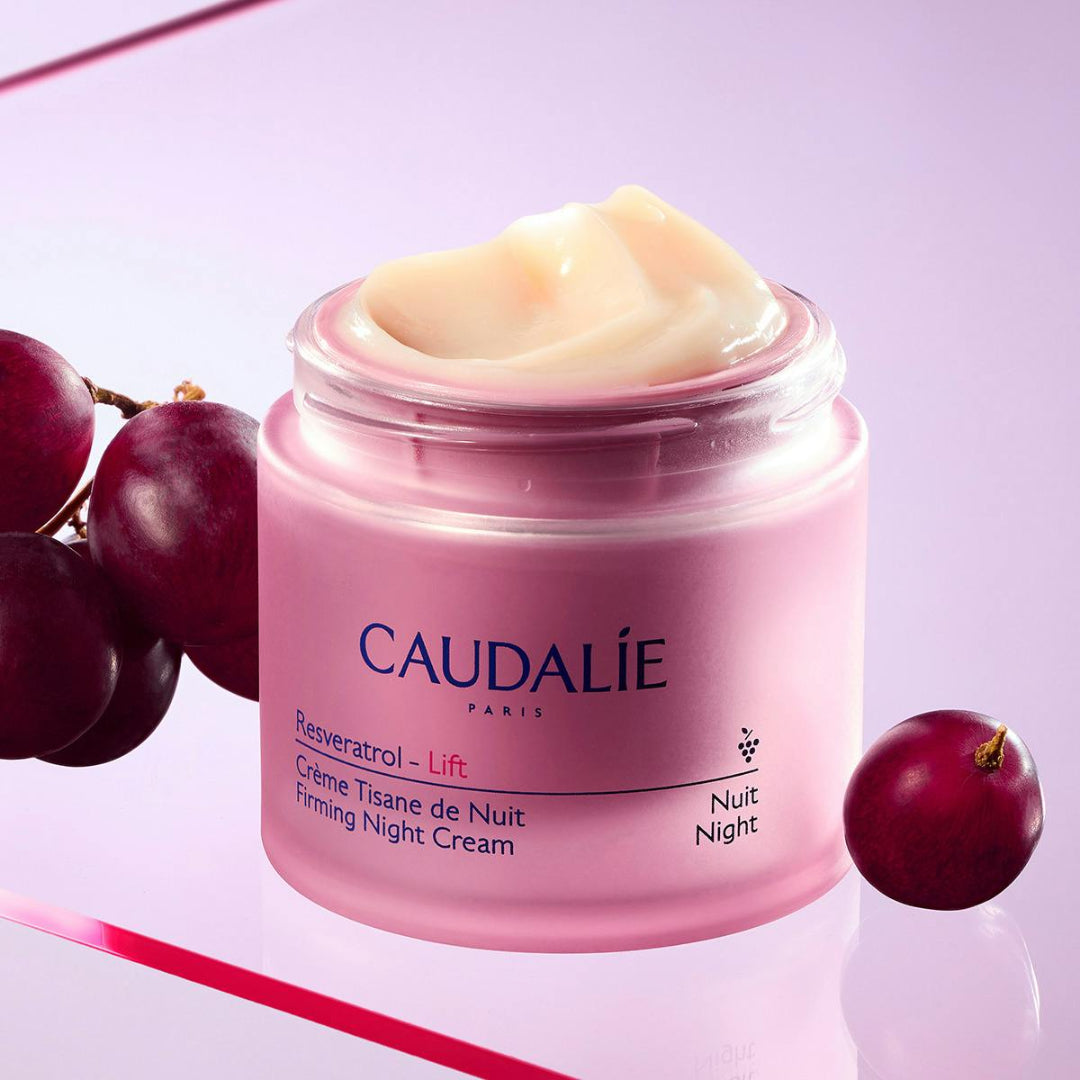 A jar of Caudalie Resveratrol - Lift Firming Night Cream with cream visible at the top, styled with red grapes on a light purple background.