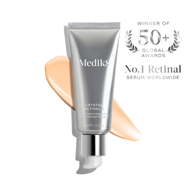 Medik8 Crystal Retinal 3 serum in a metallic silver squeeze tube with a white cap, shown with a peach-colored cream swatch behind it on a white background. To the right is a silver award badge reading 'Winner of 50+ Global Awards No.