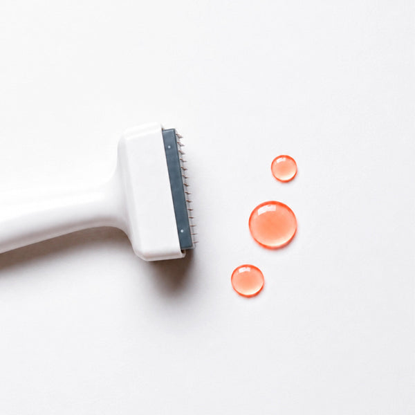 A close-up of a white pet grooming brush with a dark gray metal edge, positioned next to three clear droplets of orange liquid on a clean white background.