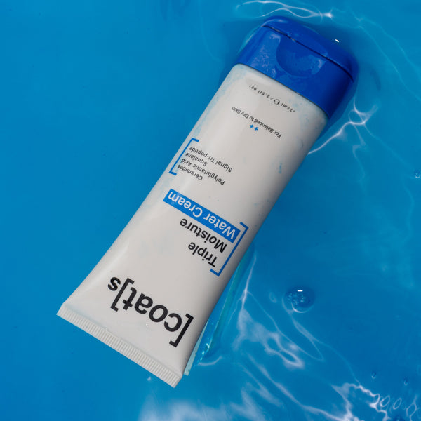 A white tube of [Coati] Triple Moisture Water Cream with a blue cap, floating on bright blue water with ripples and bubbles.