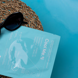 A light blue foil packet labeled 'Cool Me' and 'AIMX' lies on a woven straw hat next to black sunglasses against a bright turquoise-blue background.