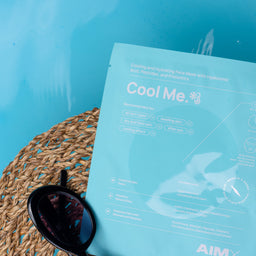 A flat-lay of a light blue cooling face mask packet labeled 'Cool Me' resting on a woven straw mat next to black sunglasses against a bright turquoise water background.
