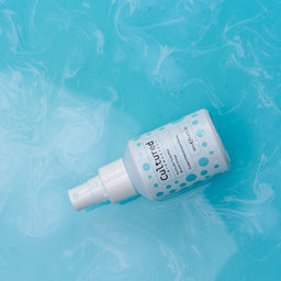 A white spray bottle labeled 'Cultivated' with blue polka dots, lying diagonally on a bright turquoise background with a marbled liquid texture.