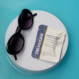 A Dr. Vegan Debitosc & Detox blister pack lies on a round white platform next to a pair of black sunglasses, all set against a bright turquoise background.