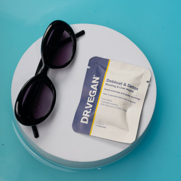 A Dr. Vegan Debitosc & Detox blister pack lies on a round white platform next to a pair of black sunglasses, all set against a bright turquoise background.