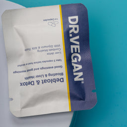 A single DR.VEGAN Debloat & Detox capsule packet, featuring a white and dark purple design with a yellow stripe, placed on a blue and white surface.