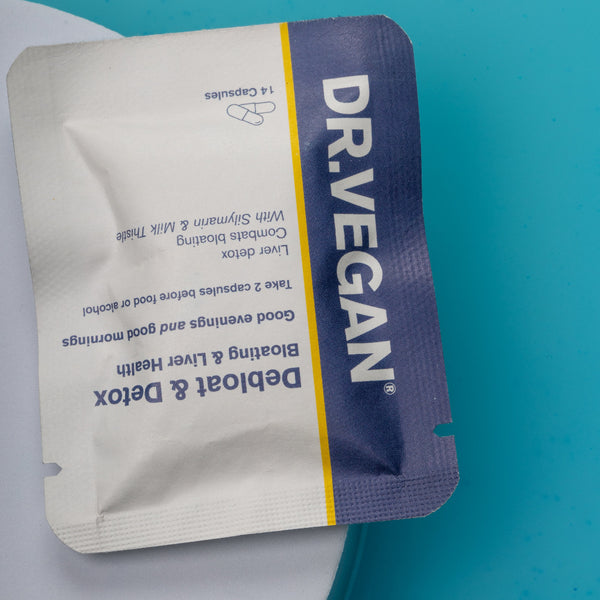 A single DR.VEGAN Debloat & Detox capsule packet, featuring a white and dark purple design with a yellow stripe, placed on a blue and white surface.