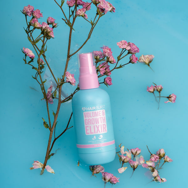 A light blue spray bottle of HairSkins Volume Growth Elixir with a pink cap, styled on a bright blue background with pink flowers.