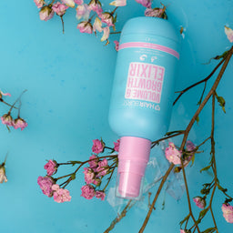 A light blue bottle of Hair & Skin Elixer Volume 30 hair growth serum with a pink spray nozzle, styled on a bright blue background with pink dried flowers.