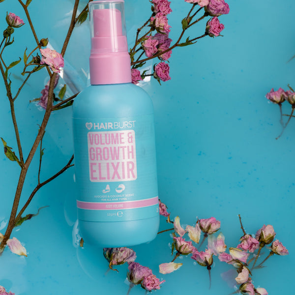 A bottle of Hairburst Volume & Growth Elixir spray, featuring a light blue body and pink pump, styled with pink flowers on a bright blue background.