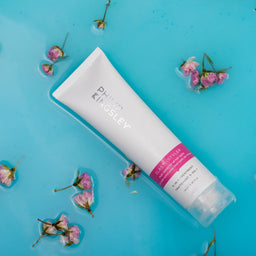 Philips Kingsley Purifying Cleansing Foam in a white squeeze tube with a pink label band, presented diagonally on a bright blue background with decorative pink flower petals.