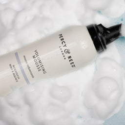 Percy by Reed Volumising Mousse body wash in a white bottle with a black pump, surrounded by thick white foam on a light blue background.