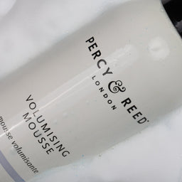 A close-up of a Percy & Reed Volumising Mousse bottle partially submerged in white foam, showing the white bottle with black text and a black cap.