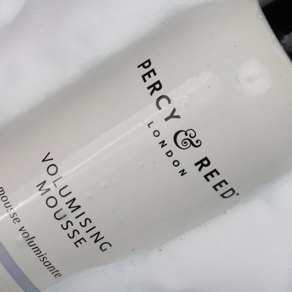 A close-up of a Percy & Reed Volumising Mousse bottle partially submerged in white foam, showing the white bottle with black text and a black cap.