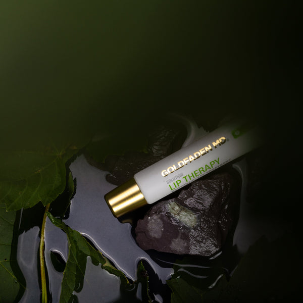 A lip therapy rollerball bottle labeled 'GOLDEN MOON LIP THERAPY' resting on dark water with green leaves and rocks in the background.