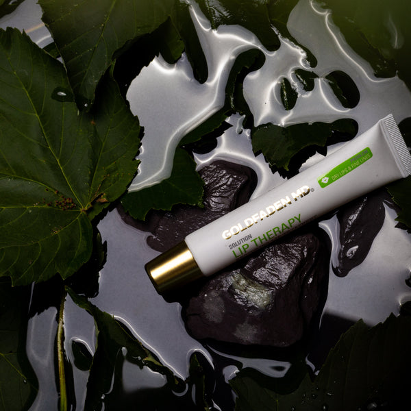 A white tube of HIG THERAPY skincare cream with a gold cap and green accents, resting on wet dark rocks surrounded by green leaves.