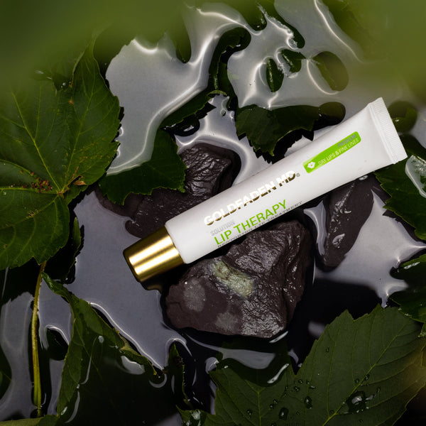 A white lip therapy cream tube with a gold cap and green accents, resting on wet dark rocks surrounded by green leaves.