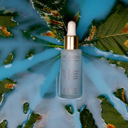 A bottle of Maken Anti-Aging Radiant Serum with a gold cap and white dropper, centered on a stylized background of blue water, green leaves, and dried orange leaves.