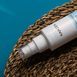 A white cylindrical spray bottle labeled KAVZEN Haircare Hair Reviver Spray, resting diagonally on a brown woven straw mat against a bright blue background.