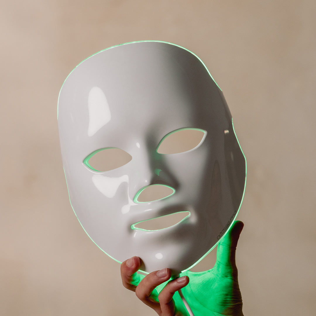 White LED face mask held by a hand wearing a green glove against a beige background