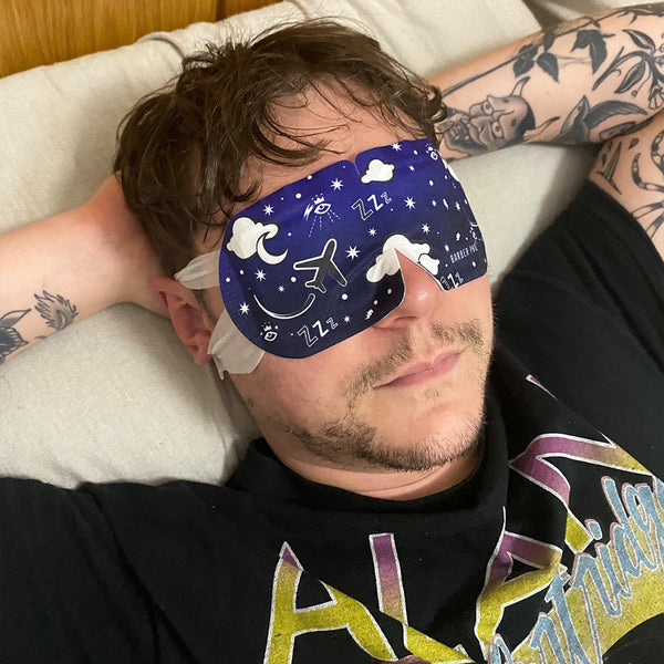 Man wearing a blue sleep mask with star and moon designs, lying on a pillow.
