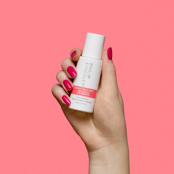 A hand with bright pink manicured nails holds a white bottle of Philip Ketsing Bond Builder Bond Enhancing Treatment against a solid pink background.