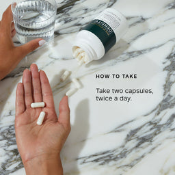 Hand holding two capsules with a bottle and glass of water on a marble surface, featuring text on how to take the capsules.