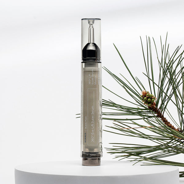 A clear cylindrical cosmetic tube with a black top and beige label stands upright on a white pedestal, with pine branches arranged along the right side against a white background.