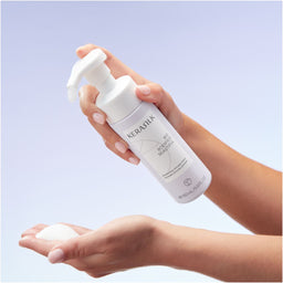 Hand holding a Keralux bottle with a pump, showing product on palm against a light purple background