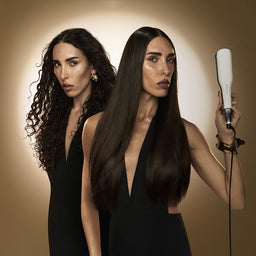 Two women with long hair, one holding a hair straightener against a brown background